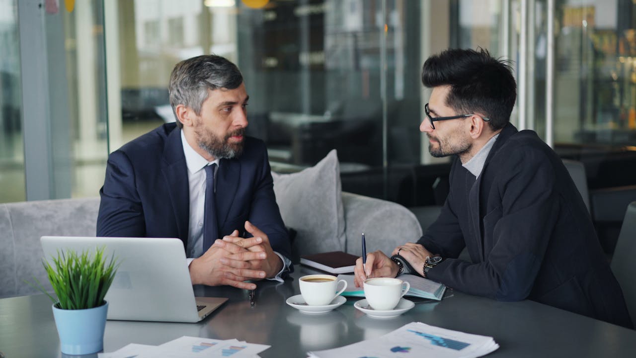 Two men in business attire sitting at a table with laptops